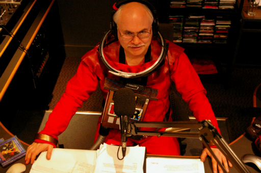Colour photo of bald man with glasses wearing a spacesuit at a radio mixing desk with microphone