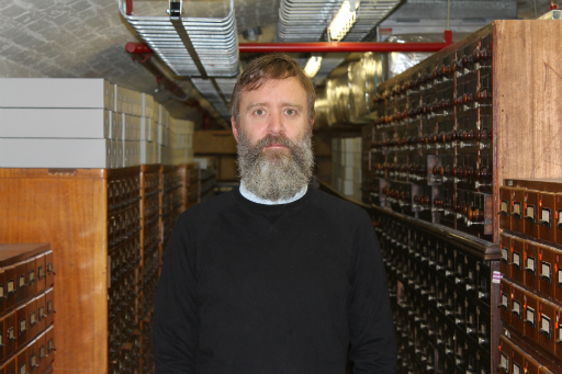 Portrait of bearded man wearing black jumper standing in a basement in front of wooden catalogue drawers