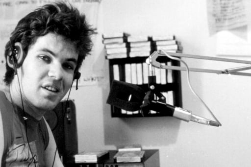 Black and white photo of young man wearing headphones and Pulp T-shirt at radio mixing desk with microphone and turntable