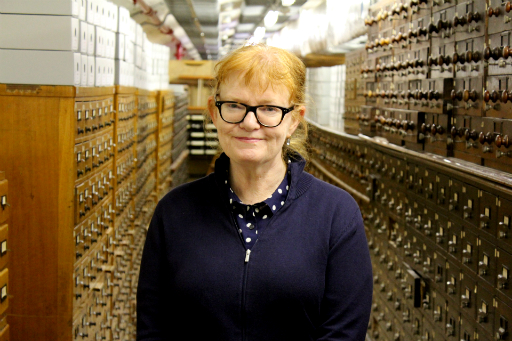 Portrait of woman wearing blue polka dot shirt and blue cardigan standing in a basement in front of wooden catalogue drawers