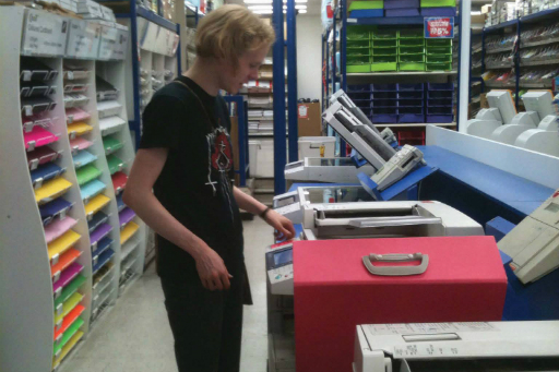 Colour photo of man with blond hair wearing a black T-shirt standing at a photocopier in a stationery store with equipment and multi-coloured paper