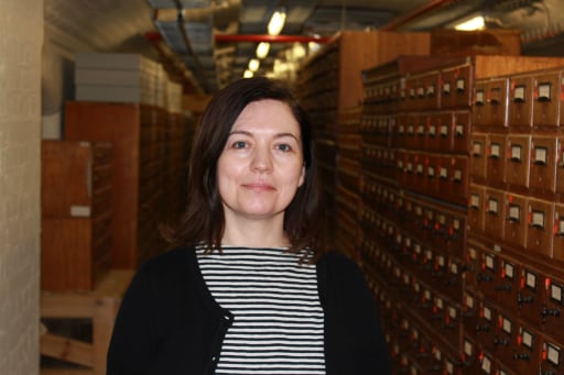 Portrait of Laura MacFarlane standing in a basement in front of wooden catalogue drawers