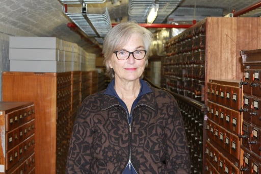 Portrait of woman wearing glasses and brown shirt standing in a basement in front of wooden catalogue drawers