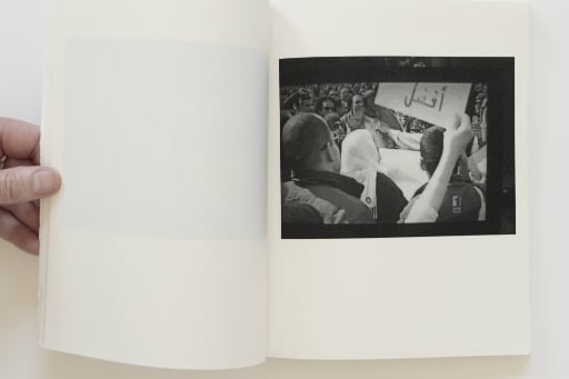 White pages of a book held open with hand, blank page on the left, the right-hand page with black and white photo of crowd of demonstrating men and women wearing headscarves, waving placards with Arabic writing