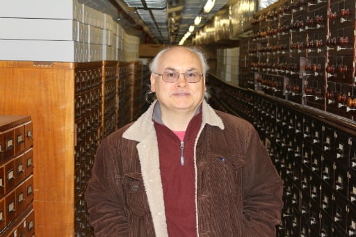 Portrait of man wearing glasses, red jumper and brown jacket standing in a basement in front of wooden catalogue drawers
