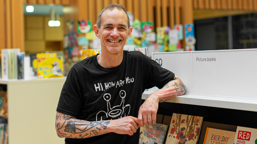 Portrait of Andy Griffiths. He is smiling and wearing a black t-shirt that says 'Hi, how are you', and is leaning against a bookshelf of picture books at the Pauline Gandel Children's Quarter at State Library Victoria.
