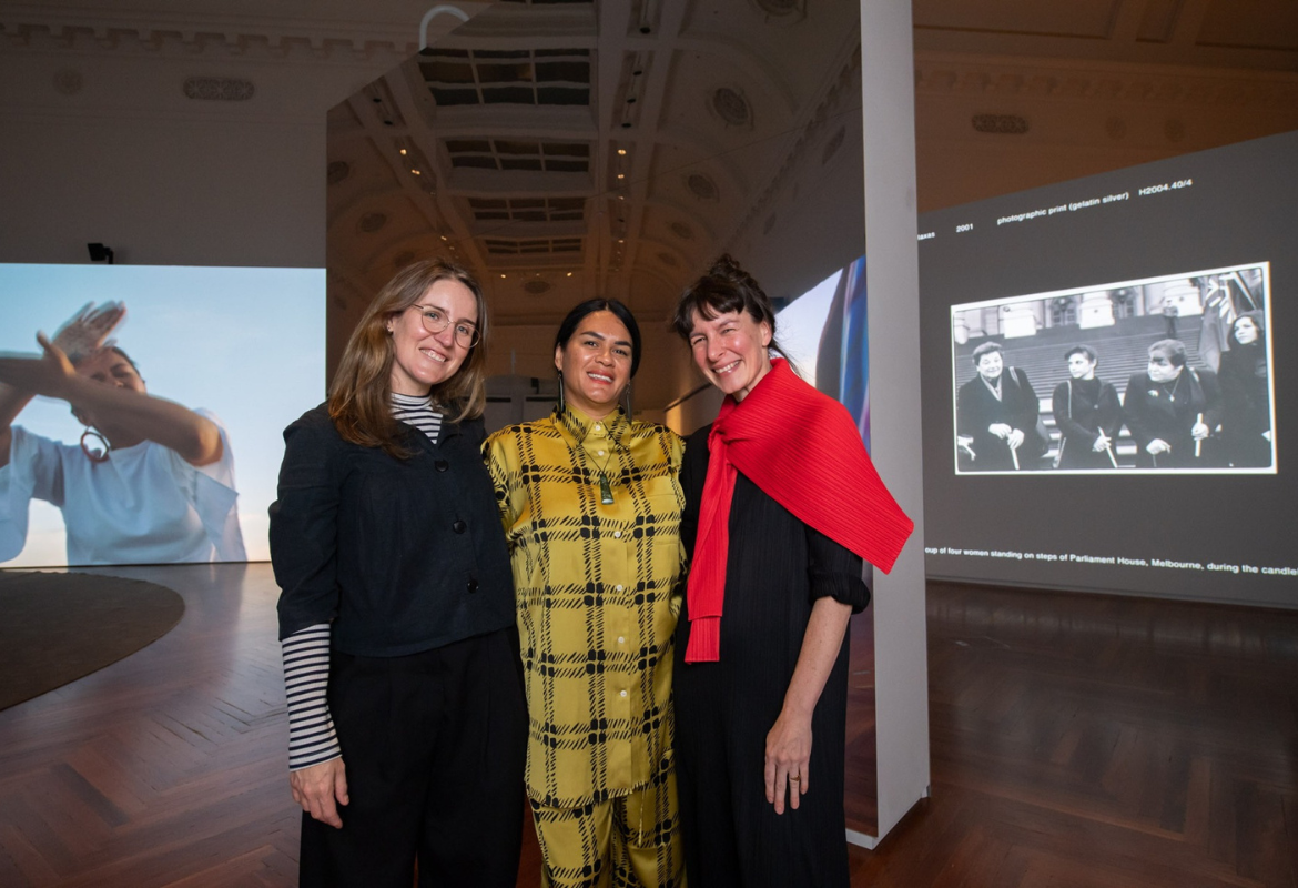 (L-R)Curators Linda Short, Jade Hadfield and Kate Rhodes standing together in the MIRROR exhibition space at State Library Victoria. They are facing the camera, with two projection screens visible in the background.