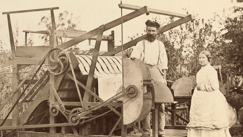 A 19th century man and woman stand behind a large piece of farm equipment