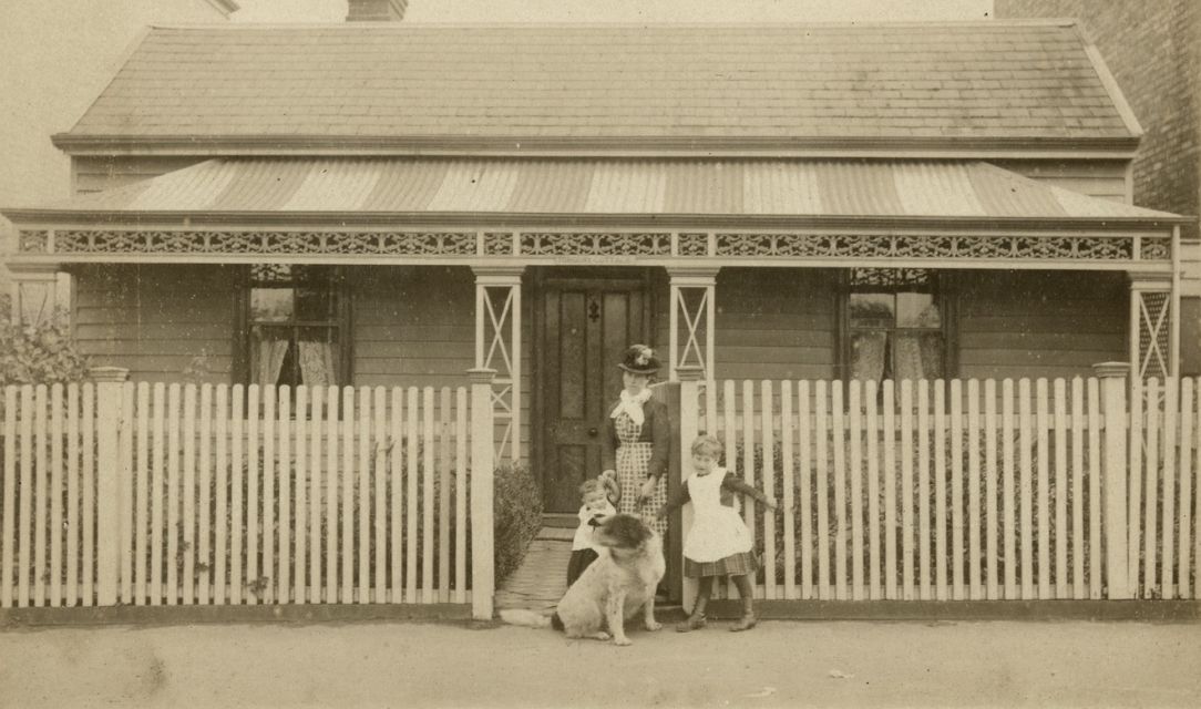 A woman standing at the gate of a weatherboard house holding the hand of a toddler with a little girl on her left, dog in front. House has a sign attached to porch roof over door "Torquay Cottage". Larger buildings on either side.