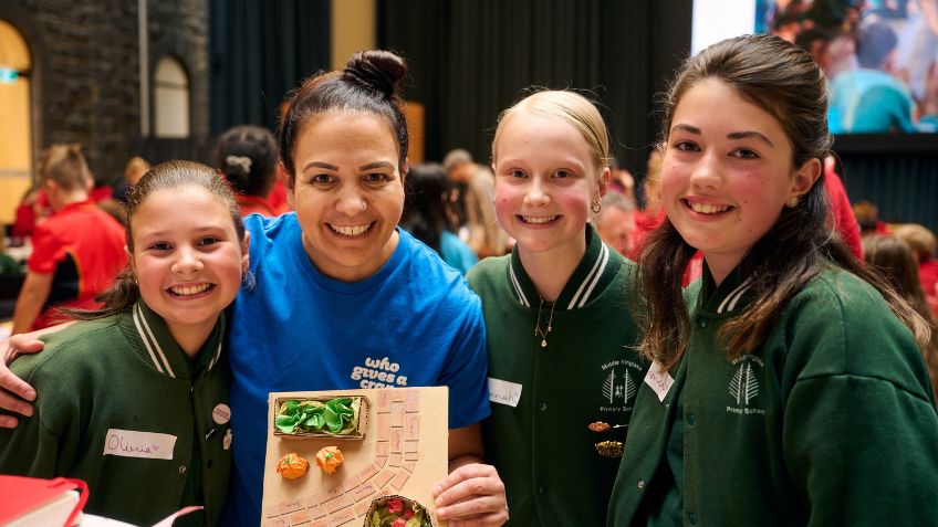 Three young students in dark green bomber jackets, smiling at the camera with an adult in a bright blue t-shirt