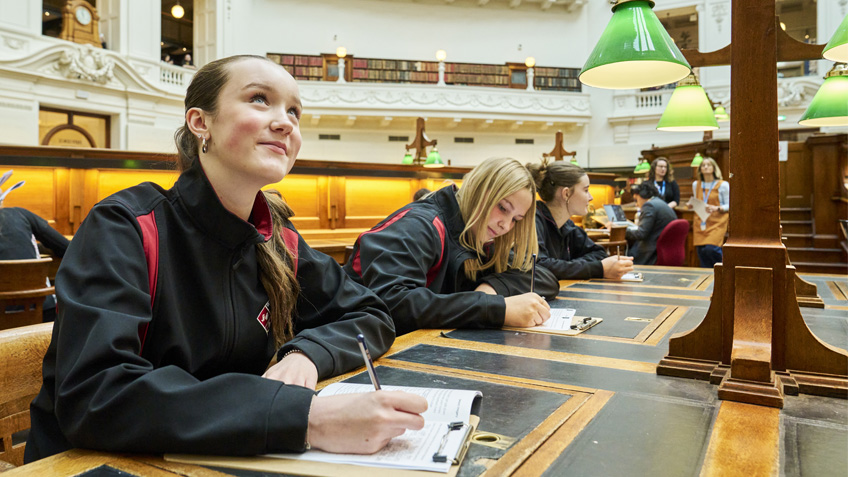 Teenager writing on notepad at a desk in the Dome. She is gazing up at the roof.