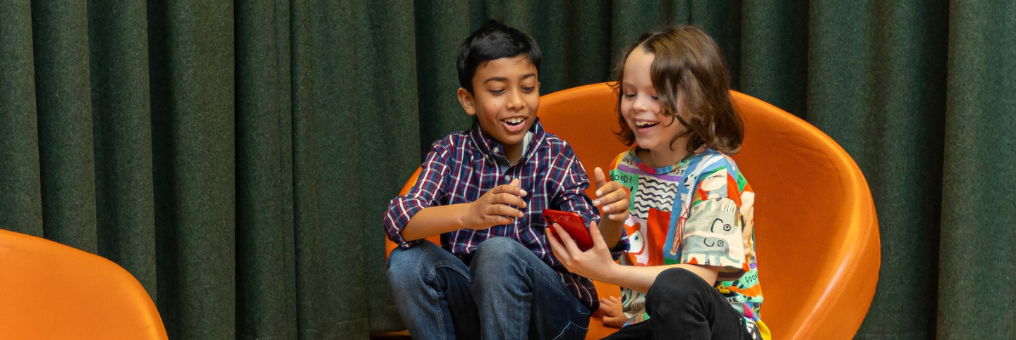 Image of two children sitting in an orange chair in the library. They are looking at a mobile phone screen.