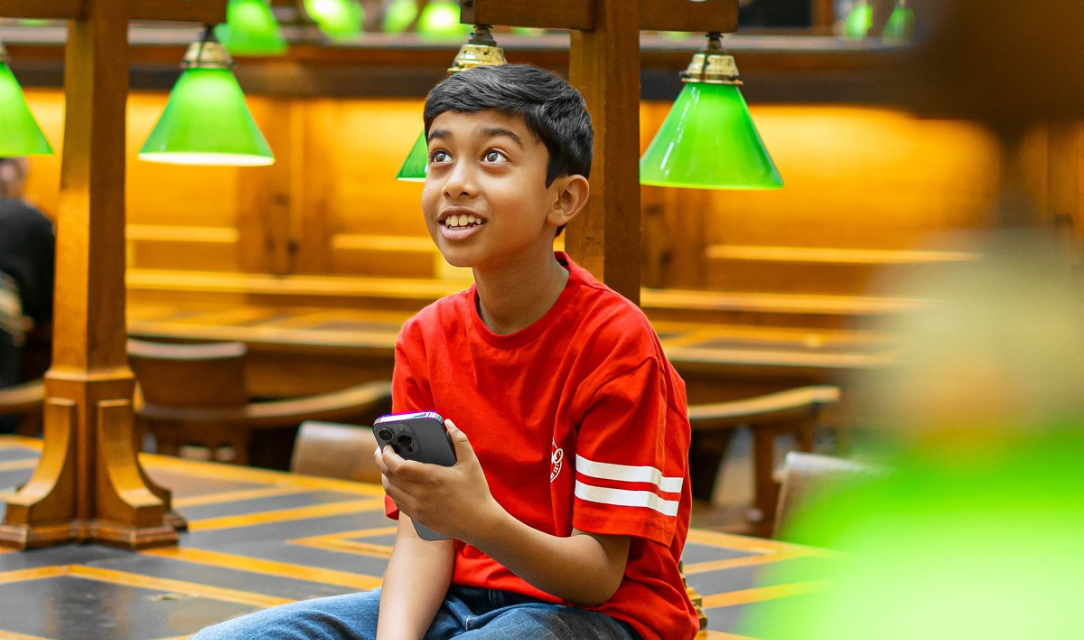 Image of a child in a red t-shirt in the La Trobe Reading Room. He is sitting on a table with a mobile phone in his hand.