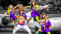 Two adults and 4 young children, all wearing tie dyed shirts and smiling at the camera. Doing an acrobat pose