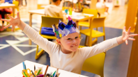 A young child sitting in the Library smiling while wearing a blue paper crown and having her hands wide spread