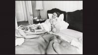 Black and white portrait of smiling Malcolm Fraser in bed speaking on telephone