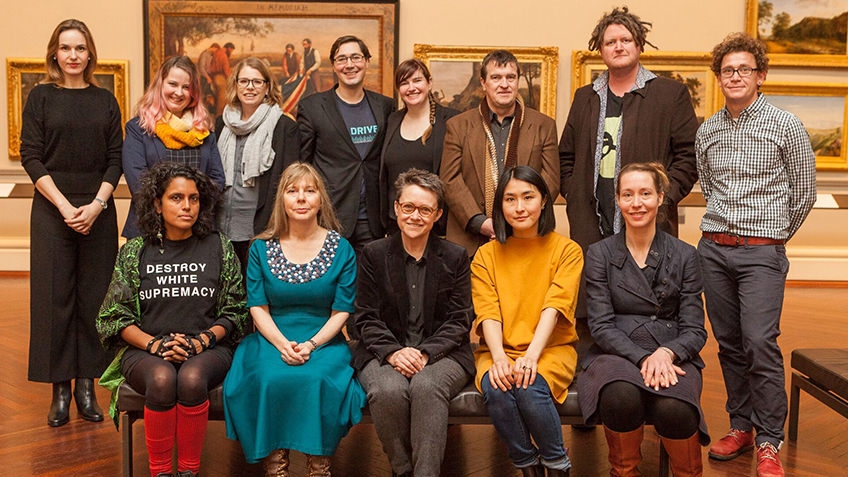 group shot of men and women seated and standing against a backdrop of classical paintings