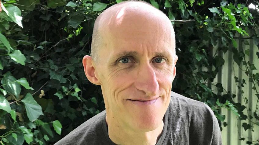 Bald man, smiling at camera in grey shirt with leaves and greenery behind him.
