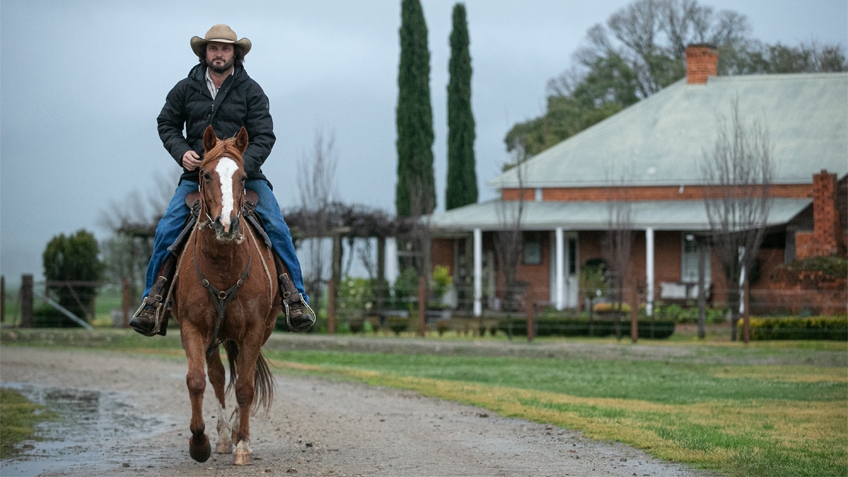 A man in a cowboy hat rides a horse with a homestead in the background