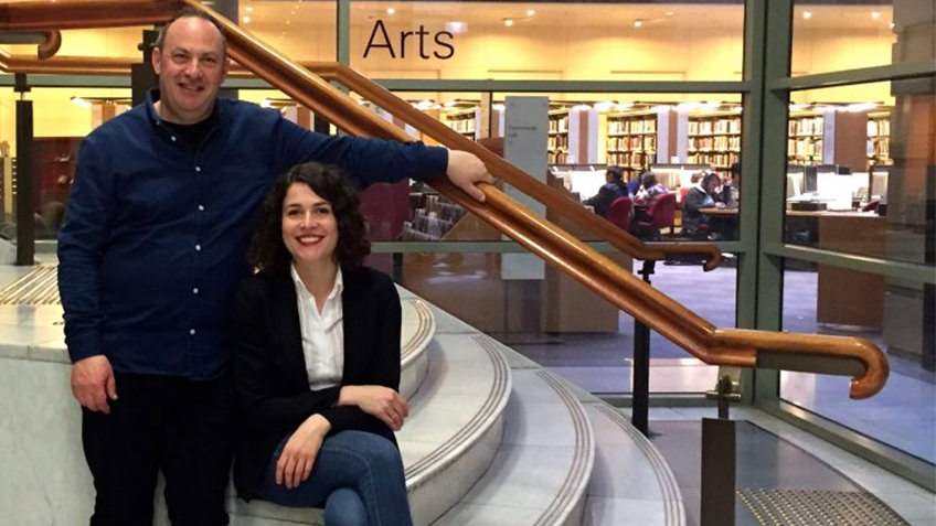 man in blue and seated woman in jeans on staircase in foyer, in background students at desks in Arts library
