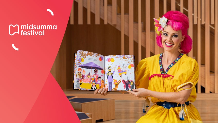 Woman with hot pink hair and yellow dress smiling at the camera, with a storybook opened and facing the camera.