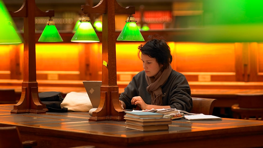 A high school student writes beneath a green desk light in a dim room