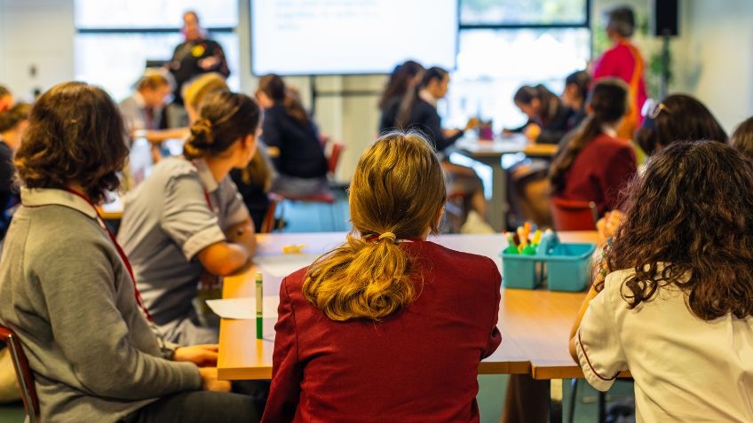 In front of you are a large group of secondary school students in a classroom, facing a screen, mid-lesson