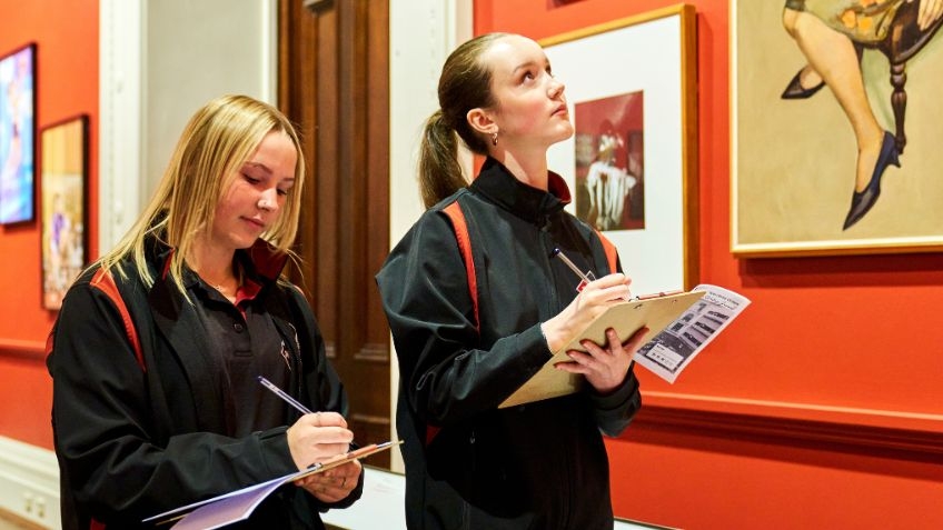 Two students in a red gallery room filled with portraits. In their hands are note pads. They are looking at the paintings and writing.