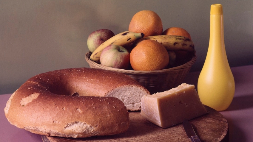 A close-up photograph of a loaf of bread, a basket of fruit and a wedge of cheese