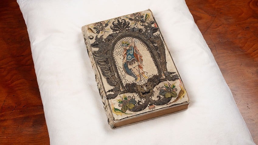 Image of a rare book with embroidered binding thought to belong to Queen Henrietta Maria, wife of King Charles I, from the John Emmerson Collection. The binding is ornate and detailed, and can be found across the entire book. The volume sits on a white pillow placed on a brown table.