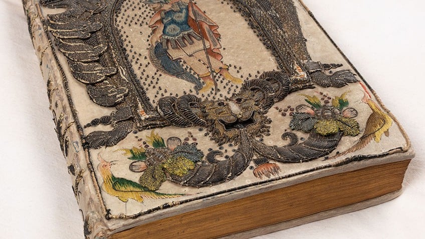 Close up image of a rare book with embroidered binding thought to belong to Queen Henrietta Maria, wife of King Charles I, from the John Emmerson Collection. The binding is ornate and detailed, and can be found across the entire book. The volume is placed on a white pillow.