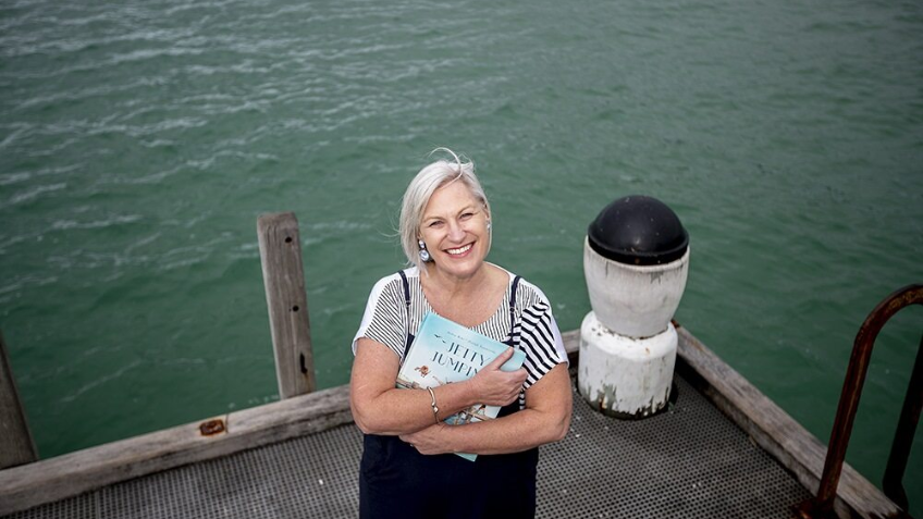 Older woman with short, cropped hair and striped shirt on a jetty with water in the background. She is holding a picture book called Jetty Jumping