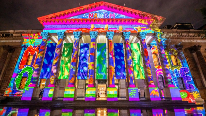 A grand building with large, Ancient Greek-style pillars. Projected onto the building are colourful graphics