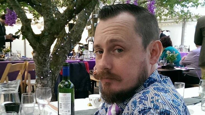 man with beard photographed under a tree with purple flowers and a table set with wine and wineglasses