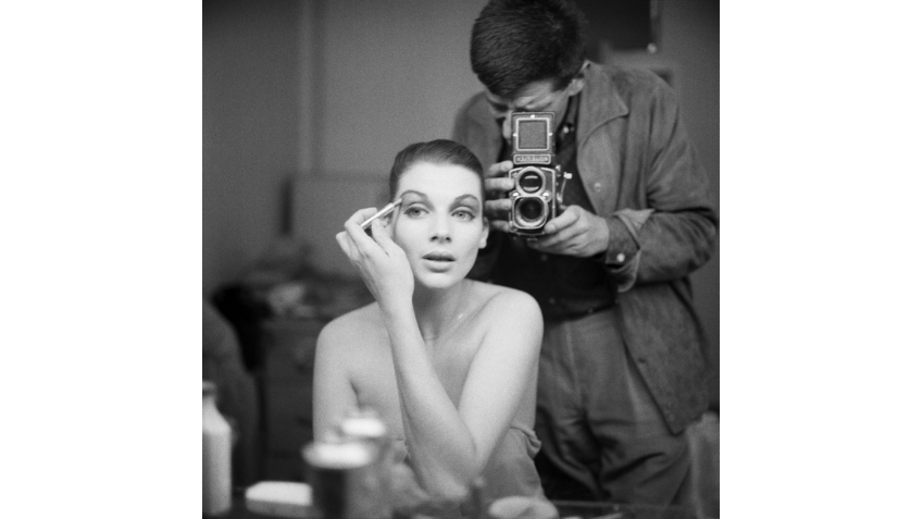 Woman applying makeup in a mirror with a man taking her photo from behind