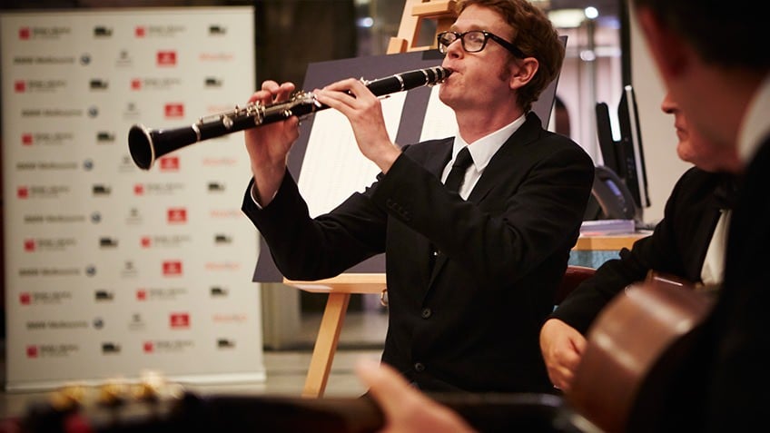 A man wearing glasses and a tuxedo plays clarinet to an audience