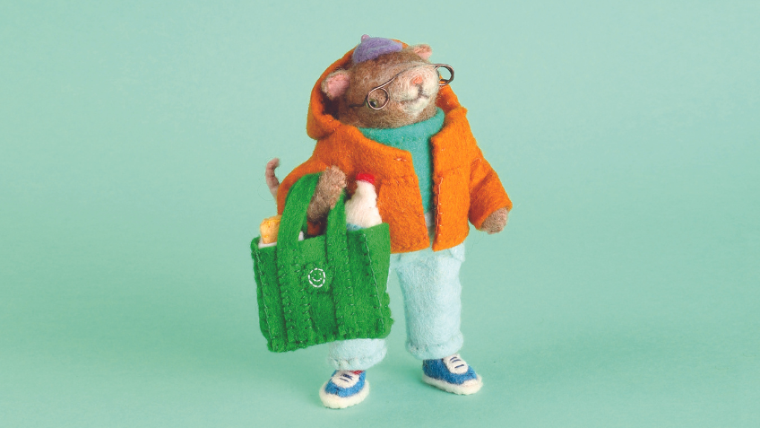 A furry creature hand-crafted by Soft Stories wearing an orange jacket and holding a green shopping bag