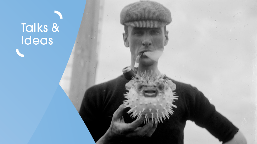 A black and white image of a man standing, half-length, wearing cloth cap and smoking pipe, holding a puffed up puffer fish.