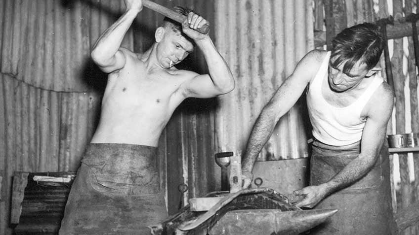 Two blacksmiths working with hammers and an anvil to repair a motor part during World War II