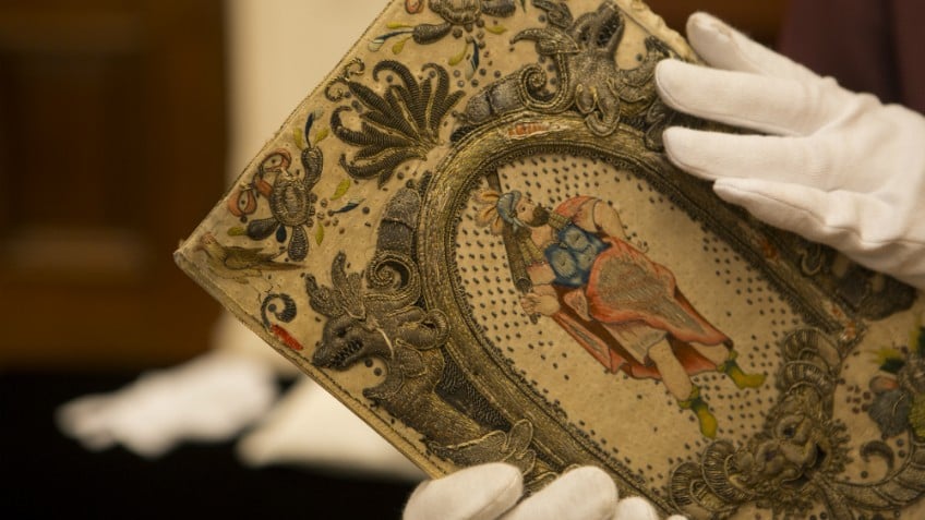 Colour photo of gloved hands holding colourfully embroidered historic binding thought to belong to Queen Henrietta Maria, wife of King Charles I, c1630