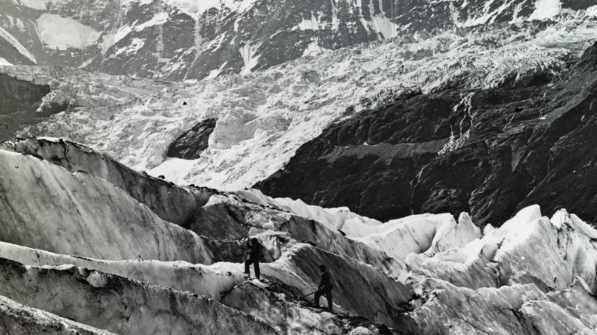 Black and white photo of small group of mountaineers ascending snow covered alps, possibly in Switzerland, carrying walking sticks or mountain poles