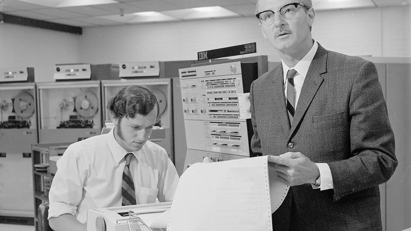 B&W photo of two men in 1970s attire standing next to an IBM computer, a dot matrix printer and reel-to-reel machines