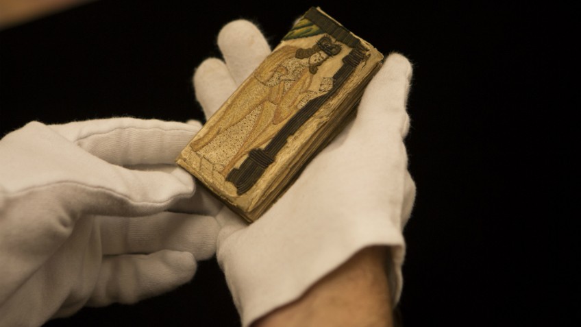 Colour photo of gloved hands holding the spiritual memoir of King Charles I, Eikon Basilike, London, 1649, featuring an embroidered image of Charles I