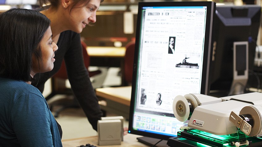 Colour photo of person using a microfiche and being helped by staff at State Library Victoria