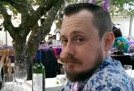 man with beard photographed under a tree with purple flowers and a table set with wine and wineglasses