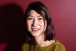 portrait of young woman with short dark hair wearing a yellow jumper against red background