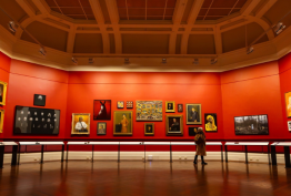 Wide angle view of the North Rotunda at State Library Victoria. The room is painted red and features artwork from the State Collection hung salon-style on its walls. There is a visitor viewing the artwork on the right side of the room.