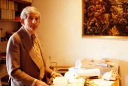 older man standing at desk in art gallery office with papers and telephone, paintings in background