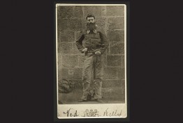 Ned Kelly in chains black background with sepia photo of bearded man in chains against bluestone wall