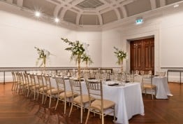Long tables are laid with white cloths, crystal glasses and flowers in an ornate heritage room